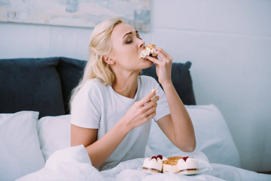 stressed woman in pajamas eating sweet cake in bed alone